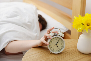 Woman turning off alarm clock on bed at home, selective focus