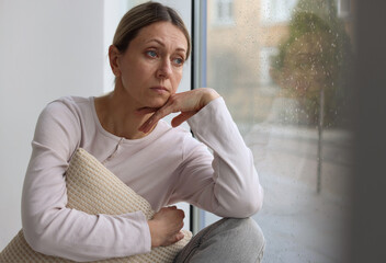 Depressed woman looking out window at home, space for text