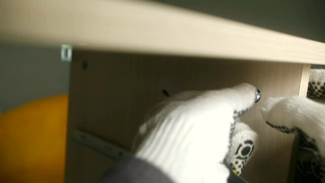 Professional furniture assembler wearing protective gloves carefully tightening screws into wooden shelf components using precise screwdriver during workshop assembly process