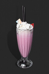 Studio-style photograph: tall glass of soft pink strawberry milkshake, topped with fluffy whipped cream, colorful marshmallow, and a slice of strawberry. Two black straws are placed in the drink. 