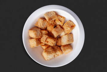 Top-down photo: crispy golden croutons cut into cubes, served in a white ceramic bowl on a black background. 