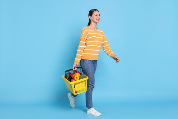 Smiling woman with shopping basket full of products waving hello on light blue background