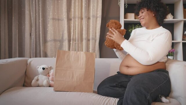 Charming pregnant woman playing with cute teddy bears, toys for future baby. African american black lady resting alone in cozy apartment, healthy pregnancy, good mood and positivity, happy parenthood