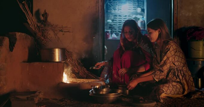 Indian villager mom with teen age female girl prepare food using earthen clay mud stove indoor house Asian young adult woman lady wear red saree cooking on open fire flam inside kitchen home desi life