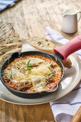 Shakshuka with baked eggs in tomato sauce, topped with melted cheese and basil, served hot in a cast iron skillet with rustic bread on the side.
