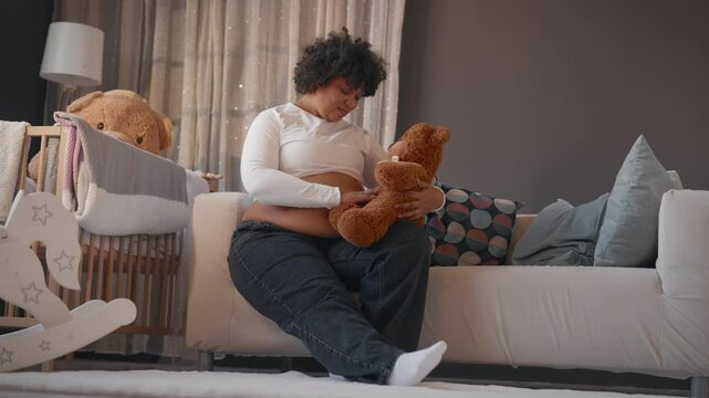 African american woman sitting alone in cozy apartment, stroking pregnant belly. Lady looking at teddy bear, waiting for her first baby, eighth month of pregnancy, firstborn in family, motherhood