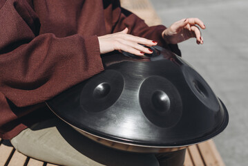 young caucasian woman playing black handpan with hands, ethnic metal drum instrument, sits on wooden bench of enbankment, beautiful relaxing music for meditation, close-up view of hands