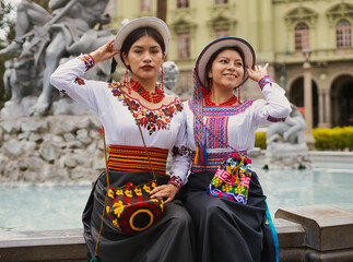 Two women in traditional, colorful attire pose by a historic fountain. Their outfits showcase intricate embroidery