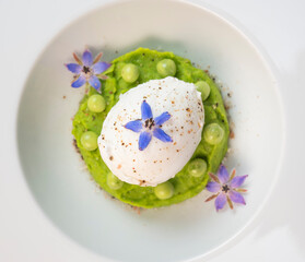 A fresh pea puree is beautifully plated, featuring a poached egg at the center and adorned with...