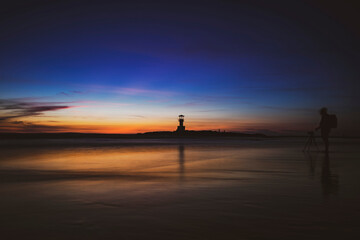 Golden hour silhouette of a lighthouse and photographer reflected on serene beach at sunset, capturing peaceful twilight ocean landscape magic.