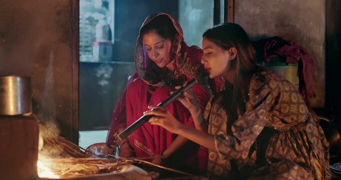 Indian villager mom with teen age female girl prepare food using earthen clay mud stove indoor house Asian young adult woman lady wear red saree cooking on open fire flam inside kitchen home desi life