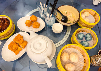 Top-down view of assorted dim sum dishes including chicken feet, shrimp balls, tofu rolls, hakau, crystal dumplings, and wontons in broth, served with tea on a round glass table.

