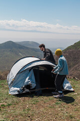 Fototapeta premium Young family camping on a hilltop with valley views. Pitching a tent, cooking outdoors, drinking coffee, and playing games. Summer adventure with full backpacking gear