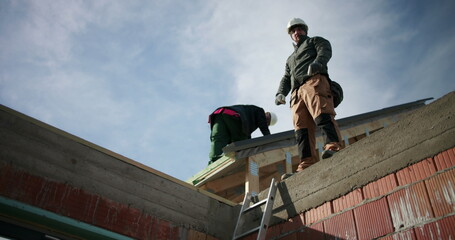Construction workers securing roof structure on ladder, focused teamwork on building site, safety gear, scaffolding, blue sky backdrop, industry professionals on a sunny day