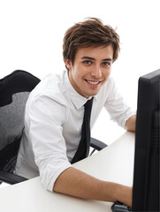Smiling young man working diligently at a modern office desk. He wears a white shirt and black tie, interacting with a computer screen while sitting in a stylish office chair.