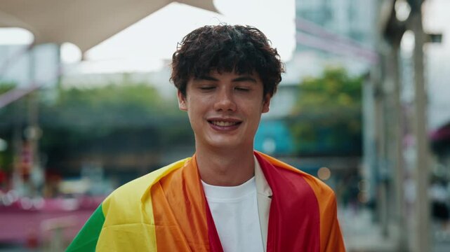 Portrait of proud young man wrapping with bright rainbow flag standing on street. Happy brunet gay shows LGBT community values outdoors. Supporting diversity and equality rights