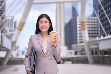 Confident young businesswoman holding a coffee cup, smiling in a modern cityscape.