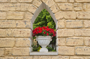 White stone vase with red petunia flowers placed in arched niche of yellow brick wall from shell...