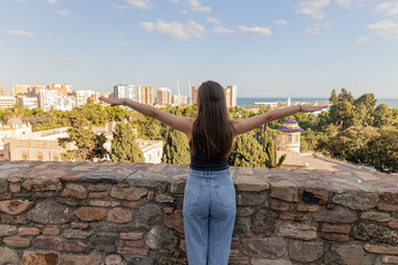 Fototapeta premium Young woman with long hair stands on stone wall, arms outstretched, enjoying panoramic view of cityscape and ocean under clear blue sky