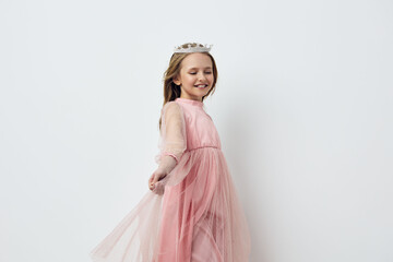 Happy young girl in a pink dress with a crown, twirling joyfully against a light backdrop,...