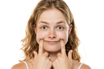 Young woman with wavy blonde hair forcing a smile by pushing her cheeks with fingers, demonstrating...