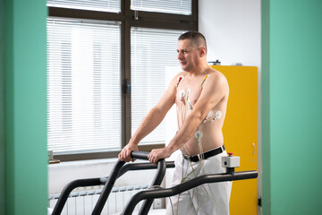 A middle-aged man is undergoing a medical stress test on a treadmill, connected to ECG electrodes for heart monitoring in a clinical environment