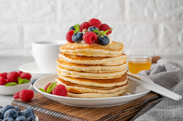 A stack of pancakes with whipped cream, fresh berries and honey on a white plate on a gray concrete...