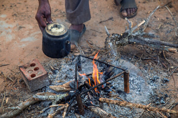 village african man cooking , tea and coffee pot on open fire in the yard at sunset, waiting for...