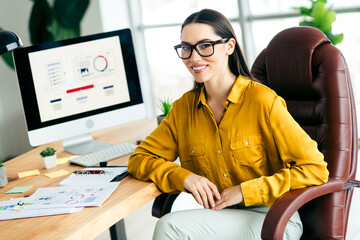 Confident young businesswoman sitting in a modern office workspace wearing a casual yellow shirt...