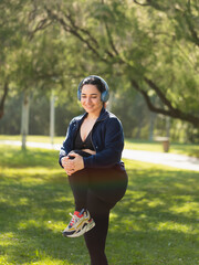 A young woman performs exercises and stretches of legs, biceps and buttocks in the morning in the park, while listening to music