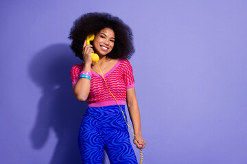 Young joyful woman speaking playfully on a vintage phone, wearing stylish retro outfit against purple background.