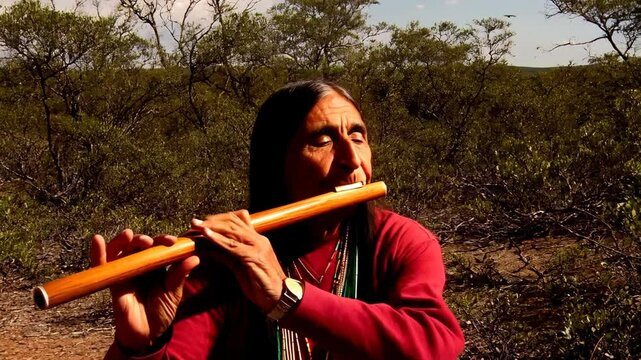 Adult indigenous man plays wooden flute outdoors surrounded by desert vegetation with eyes closed. Peaceful moment of musical expression.