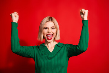 Joyful young woman raises fists in excitement against a vibrant red background wearing green sweater