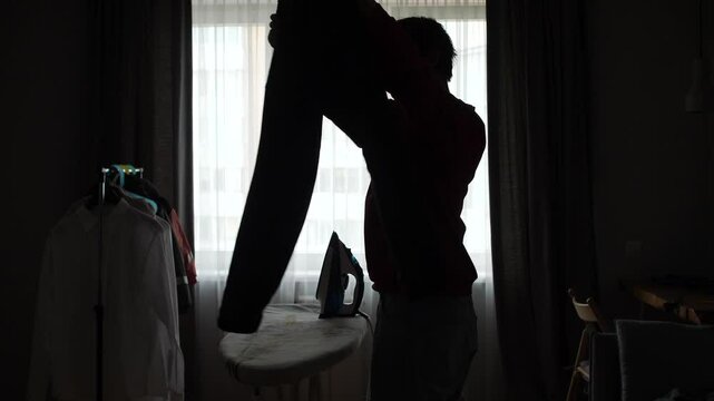 man checking trousers before ironing near window in dark room