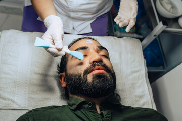 Fototapeta premium Photo of professional exfoliation, moisturizing and facial cleansing treatment for a Colombian mestizo adult male with beard where an exfoliating product is applied with a brush.