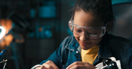 A smiling young girl sits in room in the evening with small light on, teenager is interested in fields of science, robotics, assembles her own robot, connects the electronics that power the toy.