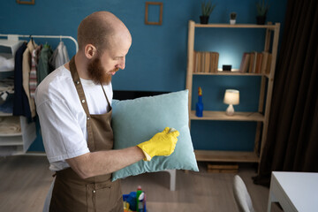 male janitor from professional cleaning service brushing pillow with brush at home interior. Housework and housekeeping