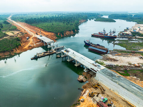 Aerial view of unfinished bridge over a river with construction machinery and boats, Eleme, Rivers, Nigeria.