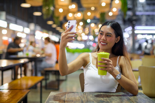 Young woman influencer using phone to take selfie picture while drinking healthy avocado smoothie drink and vlogging in cafe at night - Powered by Adobe