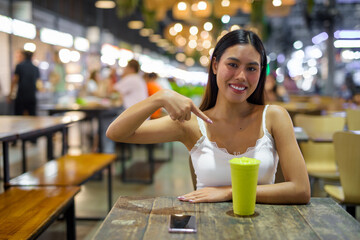 Young woman drinking healthy avocado smoothie drink in cafe at night while pointing finger to glass