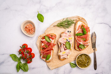 Assorted delicious sandwiches filled with thinly sliced ham, jamon or prosciutto, cream cheese, pesto and basil on a paper for breakfast arranged on white marble background, top view with copy space