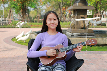 Smiling Asian preteen girl playing classic ukulele while sitting on canvas chair in the garden.