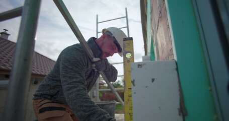 Aligning insulation panel on exterior wall with spirit level, construction worker in safety gear working on house, close-up of brick and insulation alignment