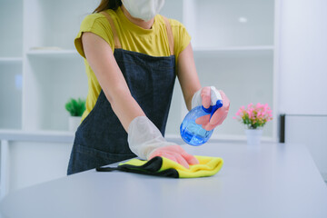 Asian woman cleans kitchen cupboard. Wipes wooden countertop with green rag. Holds spray bottle with cleaning liquid. Home interior shot. Housekeeping chore. Household hygiene concept. Woman taking