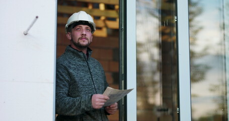 Construction worker holding blueprints, looking upward with focus, wearing hard hat, standing in doorway of unfinished building, representing planning and architectural inspection