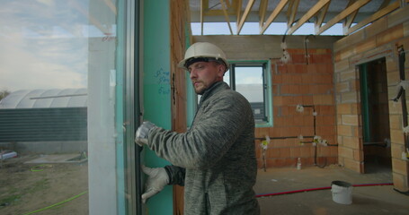 Construction worker checking window alignment and condition during building inspection, wearing hard hat and safety gloves in unfinished construction site