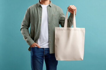 A man holds a blank beige tote bag with copy space against a teal background