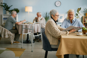 Elderly residents engaging in various activities in a cozy assisted living facility, with some reading and others chatting at tables inside a warmly lit room