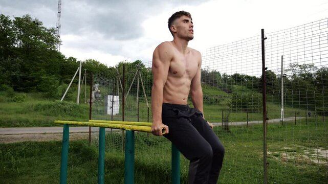 Strong guy training abdominal muscles on parallel bars at sports ground. Young shirtless sportsman exercising at nature. Sporty athlete working out at nature. Concept of active sportive lifestyle