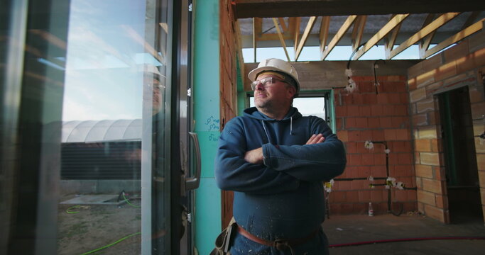 Construction worker in hard hat, arms crossed, looking outside through glass, standing inside partially completed building with brick walls